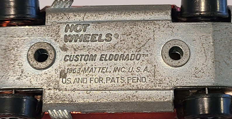 May include: Close-up of the underside of a red toy car with the text "HOT WHEELS" and "CUSTOM ELDORADO" printed on the metal chassis. The text "1968-MATTEL, INC. U.S.A." and "US AND FOR.PATS.PEND." are also visible.