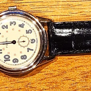 May include: A classic wristwatch with a cream-colored face and black leather band. The watch has a silver-colored case and a buckle closure. The watch face displays black numerals and hands. The band is approximately 9 inches long.