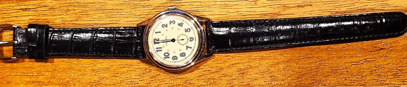 May include: A classic wristwatch with a cream-colored face and black leather band. The watch has a silver-colored case and a buckle closure. The watch face displays black numerals and hands. The band is approximately 9 inches long.