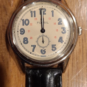 May include: A Fossil analog watch with a cream-colored face, black leather band, and silver-tone case. The watch face features black numerals, a small seconds dial, and the brand name "Fossil".