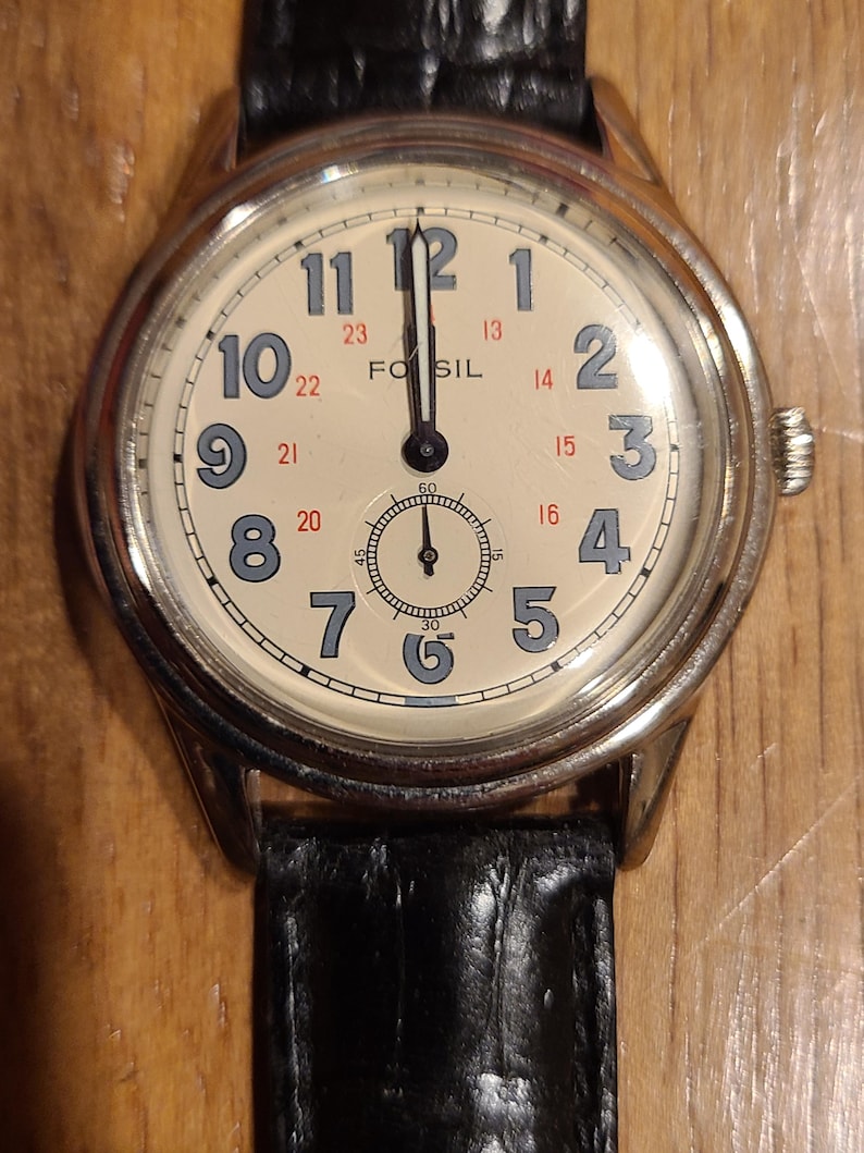 May include: A Fossil analog watch with a cream-colored face, black leather band, and silver-tone case. The watch face features black numerals, a small seconds dial, and the brand name "Fossil".