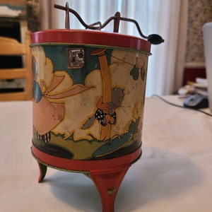 Könnte beinhalten: Ein Vintage-Blechspielzeug mit roter Basis und drei Beinen. Das Spielzeug zeigt eine farbenfrohe Illustration einer Katze, die an einer Wäscheleine mit anderen Gegenständen hängt. Das Spielzeug hat eine Kurbel oben.