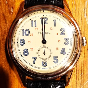 May include: A Fossil wristwatch with a cream-colored face, featuring black hands and blue numerals. The watch has a subdial at the bottom and a black leather band. The watch case is silver-toned.