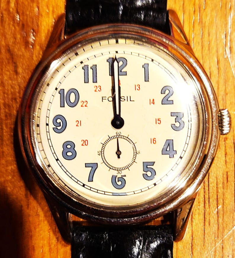 May include: A Fossil wristwatch with a cream-colored face, featuring black hands and blue numerals. The watch has a subdial at the bottom and a black leather band. The watch case is silver-toned.