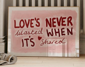 Könnte beinhalten: Ein gerahmter Kunstdruck mit dem Text "LOVE'S NEVER WASTED WHEN IT'S SHARED" in Rot auf rosa Hintergrund. Das Kunstwerk lehnt an einem weißen Heizkörper. Ein Stapel Bücher und eine Zeitschrift liegen auf dem Boden.