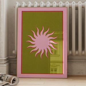 May include: A pink sun with red rays on a green background. The sun is framed in a pink and red border.