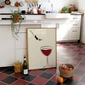 May include: A framed print featuring a diver and a wine glass with red wine. The print is leaning against a white cabinet in a kitchen with a red and black checkered floor. A bottle of wine and a jar of pasta are nearby.