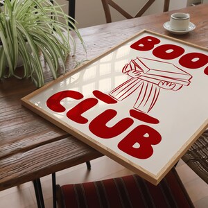 May include: A framed print featuring a red illustration of a person carrying a stack of books. The text "Book Club" is written in large red letters.