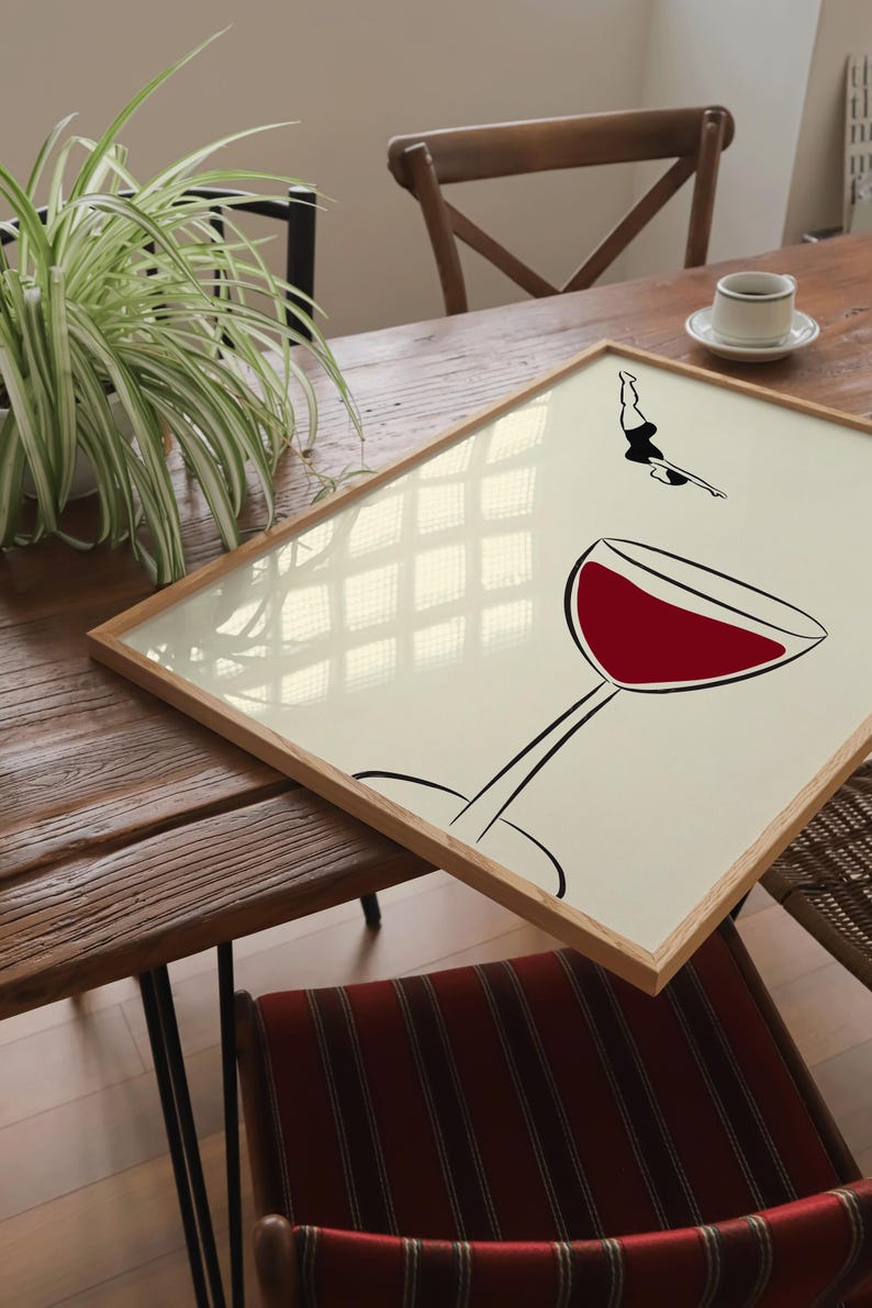 May include: A framed art print featuring a minimalist illustration of a diver and a wine glass. The wine glass is filled with red liquid. The diver is in mid-air, poised to dive. The frame is made of light-colored wood.