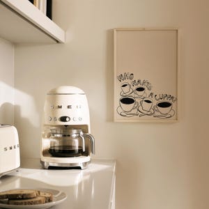 May include: A white Smeg coffee maker with a glass carafe filled with coffee. The coffee maker is on a white countertop. A framed print with the text "Who wants a cuppa?" is on the wall behind the coffee maker.
