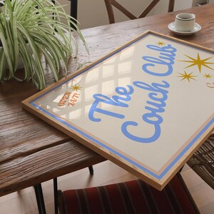 May include: A framed print with a light blue background and a white border. The print features the text "The Couch Club" in a light blue cursive font. The text is surrounded by gold stars. The print is sitting on a wooden table.