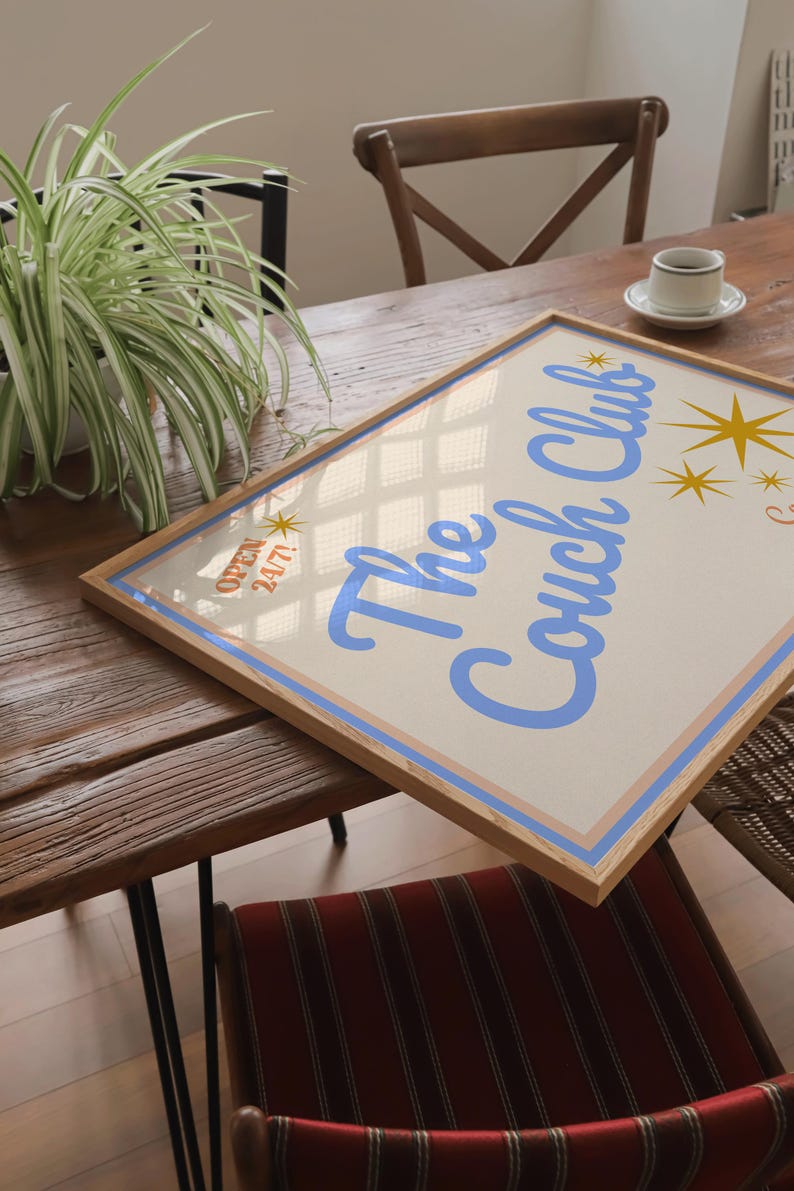 May include: A framed print with a light blue background and a white border. The print features the text "The Couch Club" in a light blue cursive font. The text is surrounded by gold stars. The print is sitting on a wooden table.
