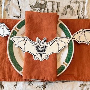 May include: A white ceramic plate with a green rim, topped with a folded burnt orange linen napkin and a die-cut paper bat decoration. The bat features a black and white ink-style illustration of a bat with wings spread wide and pointed ears.