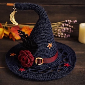 May include: A dark blue crocheted witch hat featuring a maroon ribbon band with a gold buckle, a dark red crocheted rose, black feathers, and small star appliques. A gold crescent moon charm hangs from the tip of the hat on a wooden stick.