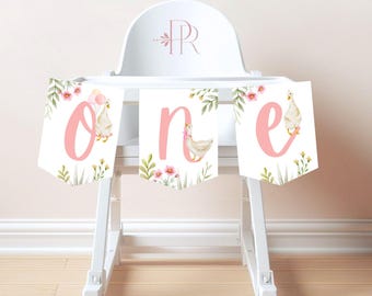 May include: A white high chair with a pink and green banner that says "one" with a duck illustration on each letter. The banner is decorated with pink flowers and green leaves.