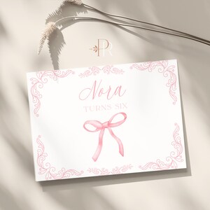 May include: A white card with pink decorative flourishes and the name "Nora" in cursive script. Below, it reads "TURNS SIX" and features a pink watercolor bow. The card is likely for a birthday celebration.