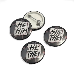 Può includere: Un set di bottoni neri con pronomi, testo bianco e illustrazione di un ratto grigio. I bottoni mostrano "HE HIM", "SHE THEY" e "HE THEY" su sfondo bianco. Un bottone è aperto, mostrando il retro della spilla.