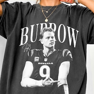 May include: A charcoal gray short-sleeve t-shirt featuring a large white graphic print of Joe Burrow in a Cincinnati Bengals jersey with the number 9. The text BURROW is printed in a bold white serif font above the player, with a signature included.