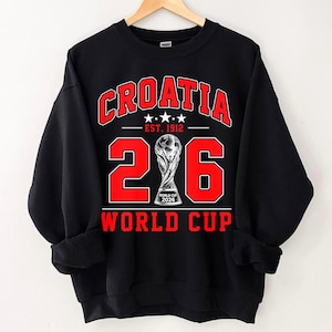 Op de afbeelding: Een zwart sweatshirt met lange mouwen en een rood-witte grafische print. De tekst luidt CROATIA, EST. 1912 en WORLD CUP. Het getal 206 wordt weergegeven met een trofee-illustratie in plaats van de nul, met het label WORLD CUP 2026.