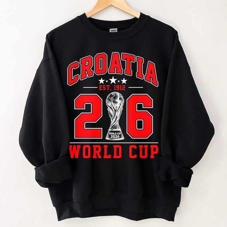 Op de afbeelding: Een zwart sweatshirt met lange mouwen en een rood-witte grafische print. De tekst luidt CROATIA, EST. 1912 en WORLD CUP. Het getal 206 wordt weergegeven met een trofee-illustratie in plaats van de nul, met het label WORLD CUP 2026.