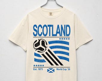 Puede incluir: Camiseta color crema con la palabra "SCOTLAND" en letras azules. Debajo, un gráfico de balón de fútbol con rayas azules y negras, y el texto "Est. 1873" y "World Cup '26".