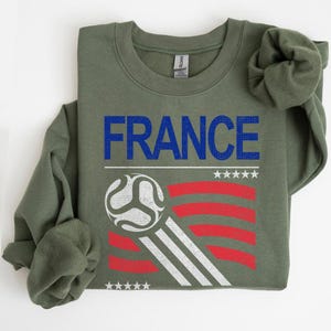 Pu&ograve; includere: Una felpa verde oliva con la scritta "FRANCE" in lettere blu. Sotto, una grafica di pallone da calcio con strisce rosse e bianche e stelle bianche. La felpa &egrave; piegata.