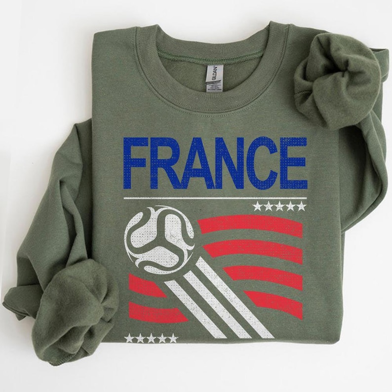 Pu&ograve; includere: Una felpa verde oliva con la scritta "FRANCE" in lettere blu. Sotto, una grafica di pallone da calcio con strisce rosse e bianche e stelle bianche. La felpa &egrave; piegata.
