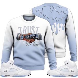 May include: White and light blue sweatshirt with a graphic of two hands shaking, a snake, and the text "TRUST No One." The back of the sweatshirt has a blue drip design. White sneakers with blue accents are also in the image.