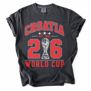 Op de afbeelding: Een antracietgrijs t-shirt met korte mouwen en een rode grafische print. De tekst luidt CROATIA, EST. 1912 en WORLD CUP. Een groot getal 26 omringt een centrale trofee-illustratie met de tekst WORLD CUP 2026 aan de voet van de trofee.