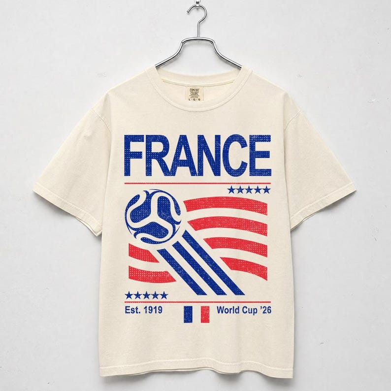 Pu&ograve; includere: T-shirt color crema con la scritta "FRANCE" in blu, sopra una grafica di pallone da calcio e strisce rosse e bianche. La maglietta presenta anche il testo "Est. 1919" e "World Cup '26".