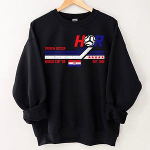 May include: Black sweatshirt with a graphic design featuring the text "HR" in red and blue, a soccer ball, and the Croatian flag. The text "CROATIA SOCCER" and "WORLD CUP '26" are also displayed. The sweatshirt has a crew neck and long sleeves.