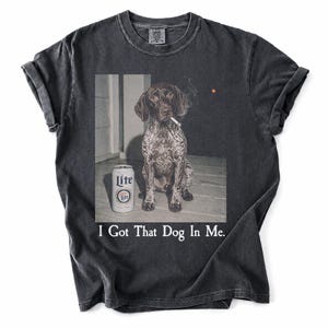 May include: A charcoal grey t-shirt featuring a graphic print of a spotted dog sitting next to a can of Lite beer. The dog has a cigarette in its mouth with a small wisp of smoke. Below the image, the text reads, I Got That Dog In Me.
