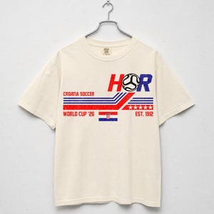 May include: Cream-colored t-shirt with a graphic design featuring the text "HOR" in red and blue, a soccer ball, and the Croatian flag. The shirt also includes "CROATIA SOCCER", "WORLD CUP '26", and "EST. 1912".