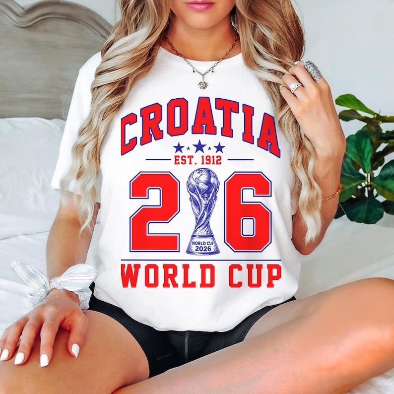 Op de afbeelding: Een wit t-shirt met korte mouwen en een rood-blauwe grafische print met de tekst CROATIA EST. 1912, het getal 2026 met een illustratie van de wereldbeker in het midden, en de tekst WORLD CUP onderaan in dikke rode letters.