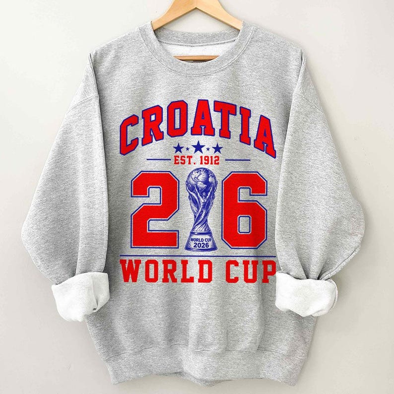 Op de afbeelding: Een grijs gem&ecirc;leerd sweatshirt met ronde hals en een rood-blauwe grafische print. De tekst luidt CROATIA EST. 1912 en WORLD CUP. Een groot getal 26 wordt weergegeven met een blauwe trofee-illustratie in het midden, inclusief WORLD CUP 2026.