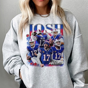 May include: A light gray crewneck sweatshirt featuring a graphic print of football player Josh Allen in a blue Buffalo Bills uniform. The design includes the text JOSH at the top and Allen at the bottom, with multiple action shots of the player.