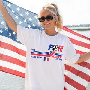 May include: White t-shirt with a graphic design featuring the word "FOR" in blue and red, a soccer ball, and the text "FRANCE SOCCER 2026 EST. 1919". The person is holding an American flag.