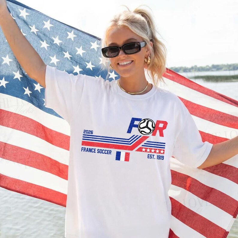 May include: White t-shirt with a graphic design featuring the word "FOR" in blue and red, a soccer ball, and the text "FRANCE SOCCER 2026 EST. 1919". The person is holding an American flag.