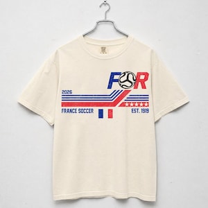 May include: Cream-colored t-shirt with a graphic design featuring the text "FOR" in blue and red, a soccer ball, and the words "FRANCE SOCCER" and "EST. 1919". The design includes blue and red stripes and the year "2026".