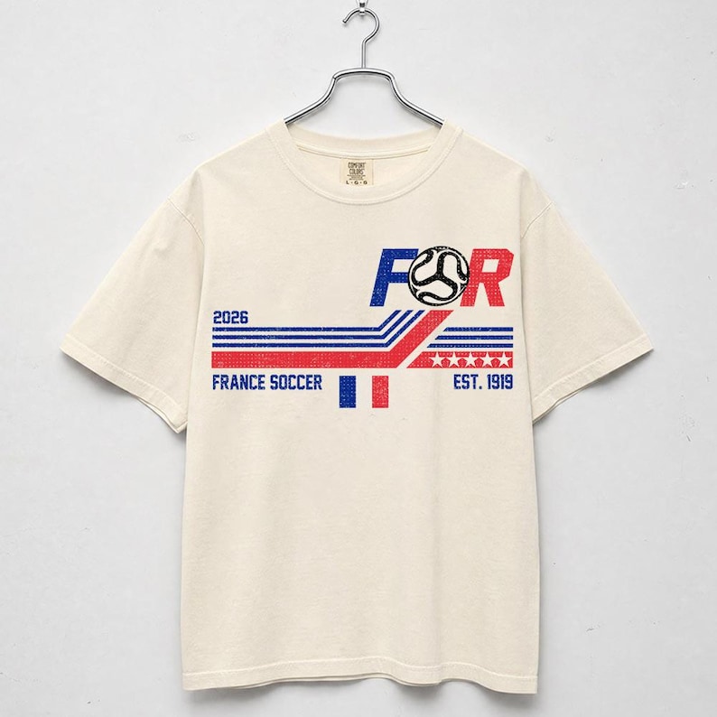 May include: Cream-colored t-shirt with a graphic design featuring the text "FOR" in blue and red, a soccer ball, and the words "FRANCE SOCCER" and "EST. 1919". The design includes blue and red stripes and the year "2026".