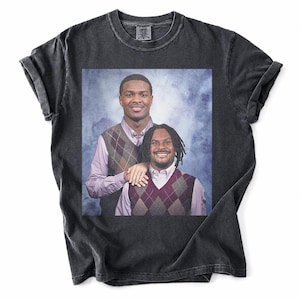 May include: A charcoal black short-sleeve t-shirt featuring a large rectangular graphic print of two smiling individuals wearing light purple button-down shirts and brown argyle sweater vests against a blue mottled studio backdrop.