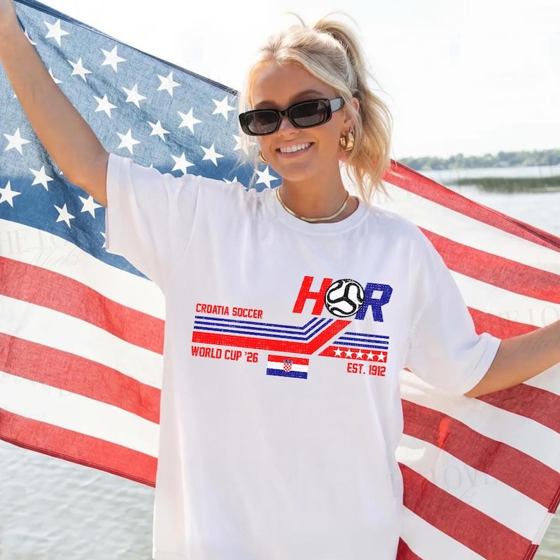 May include: White t-shirt with a graphic design featuring the text "CROATIA SOCCER WORLD CUP '26" in red and blue. The design includes a soccer ball and the Croatian flag. The shirt also has the text "EST. 1912".