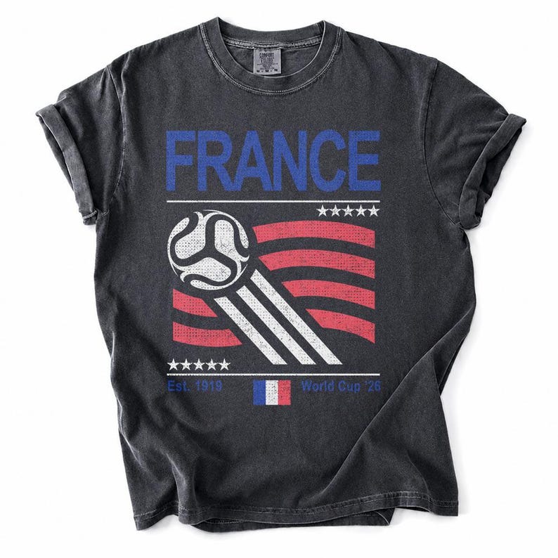 Pu&ograve; includere: T-shirt grigio scuro con un design grafico con la scritta "FRANCE" in blu, un pallone da calcio e un design a bandiera rosso, bianco e blu. La maglietta include il testo "Est. 1919" e "World Cup '26".