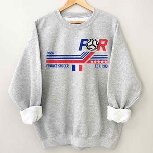 May include: A heather gray sweatshirt with a graphic design featuring the text "FOR" in blue and red, a soccer ball, and the words "FRANCE SOCCER" and "EST. 1919". The design includes red, white, and blue stripes and stars, and the year "2026".