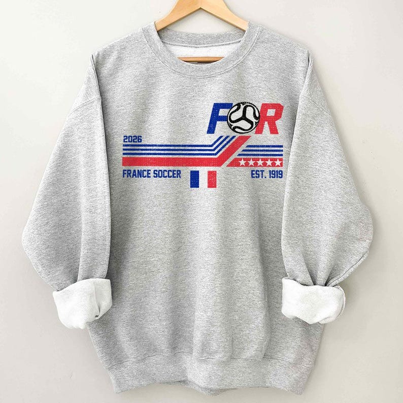 May include: A heather gray sweatshirt with a graphic design featuring the text "FOR" in blue and red, a soccer ball, and the words "FRANCE SOCCER" and "EST. 1919". The design includes red, white, and blue stripes and stars, and the year "2026".