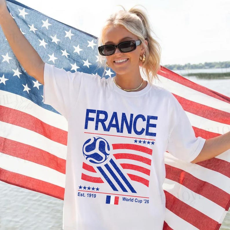 Pu&ograve; includere: Maglietta bianca con la scritta "FRANCE" in blu, una grafica di pallone da calcio e strisce rosse e blu. La maglietta presenta anche "Est. 1919" e "World Cup '26". La persona tiene in mano una bandiera americana.
