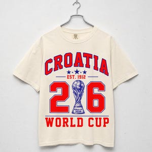 Op de afbeelding: Een cr&egrave;mekleurig t-shirt met korte mouwen en rode en blauwe grafische tekst met de tekst CROATIA EST. 1912 en WORLD CUP. Het ontwerp bevat het getal 26 met een blauwe illustratie van de World Cup 2026-trofee die de nul vervangt.