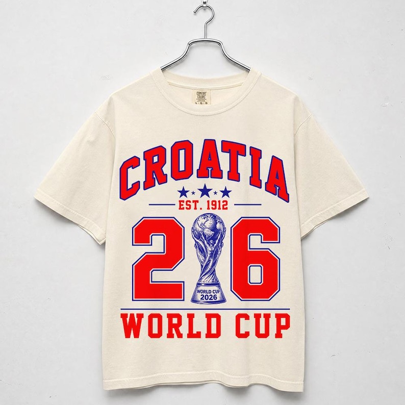 Op de afbeelding: Een cr&egrave;mekleurig t-shirt met korte mouwen en rode en blauwe grafische tekst met de tekst CROATIA EST. 1912 en WORLD CUP. Het ontwerp bevat het getal 26 met een blauwe illustratie van de World Cup 2026-trofee die de nul vervangt.