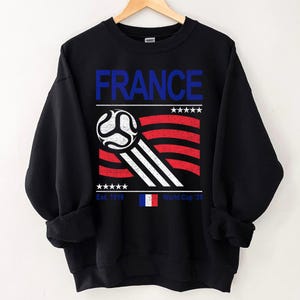 Pu&ograve; includere: Felpa nera con un design grafico con la scritta "FRANCE" in blu, un pallone da calcio e una bandiera francese stilizzata in rosso, bianco e blu. &Egrave; incluso anche il testo "Est. 1919" e "World Cup '26".