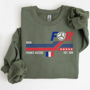 May include: Olive green sweatshirt with a graphic design for France Soccer. The design includes the text "FOR", a soccer ball, the French flag, the year "2026", and "EST. 1919". The design features red, white, and blue colors.
